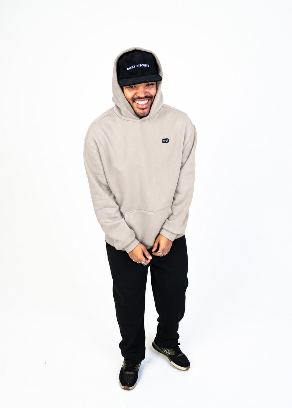A man wearing bone coloured hoodie from bro clothing with a logo badge on the wearer's left chest, the man is wearing black jogging bottoms and the photo is shot against a plain white studio background