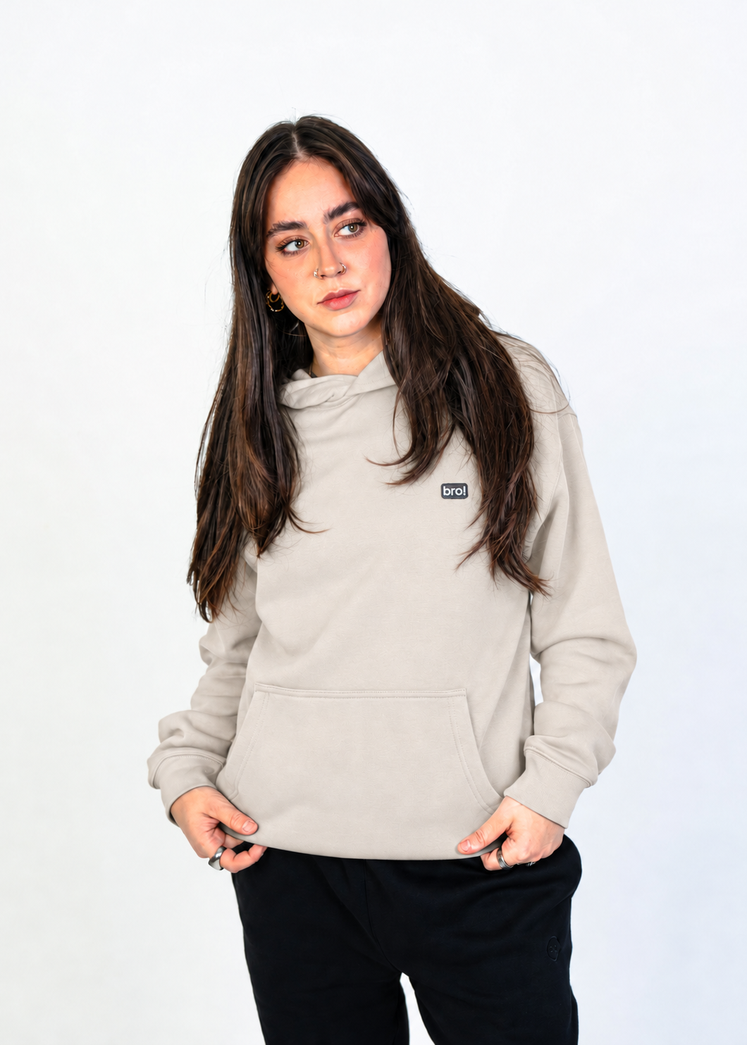 A woman with long dark brown hair wearing a light beige hooded sweatshirt with a small black logo patch on the left chest, paired with black pants, standing against a plain white studio background.